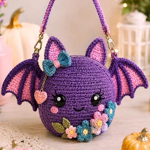 May include: A round purple crochet bag shaped like a bat with pink inner ears and wings, a blue bow, and a cluster of colorful crocheted flowers at the base. The bag features black plastic eyes, a small smile, and a dangling pink heart charm.