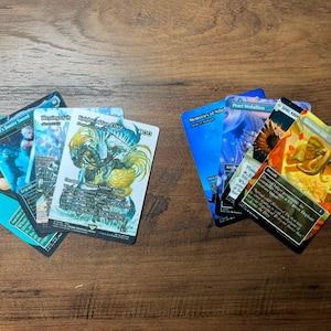Könnte beinhalten: Zwei Gruppen von Magic: The Gathering Sammelkarten auf einer Holzoberfläche. Die linke Gruppe enthält Karten wie Cloud's Buster Sword und Knight of the White Orchid. Die rechte Gruppe umfasst Karten wie Pearl Medallion und Generous Gift.