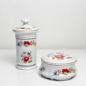 Könnte beinhalten: Zwei dekorative Keramikteile in Weiß. Eines ist ein hoher Behälter mit Deckel, das andere eine kleine, runde Dose mit Deckel. Beide sind mit einem Blumenmuster mit roten, blauen und gelben Blumen mit grünen Blättern verziert.