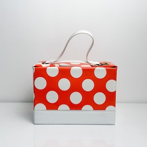 Könnte beinhalten: Quadratische Handtasche mit rotem Polka-Dot-Muster und weißen Punkten sowie einem weißen Griff. Der Boden der Tasche ist weiß. Die Tasche steht auf einer weißen Oberfläche.