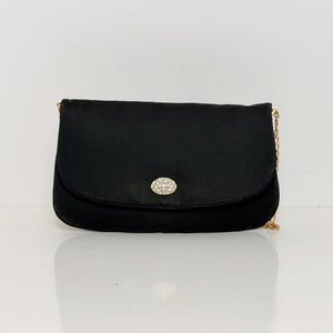 May include: A black satin clutch with a gold chain strap. The flap closure features a small, oval-shaped embellishment with sparkling rhinestones. The bag is set against a white background.