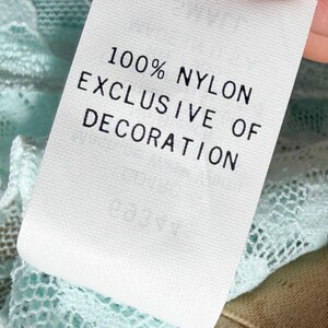 May include: A white fabric garment tag with black text that reads 100% NYLON EXCLUSIVE OF DECORATION, attached to a light mint green lace fabric with a detailed floral pattern. The tag is rectangular and positioned against the textured lace.