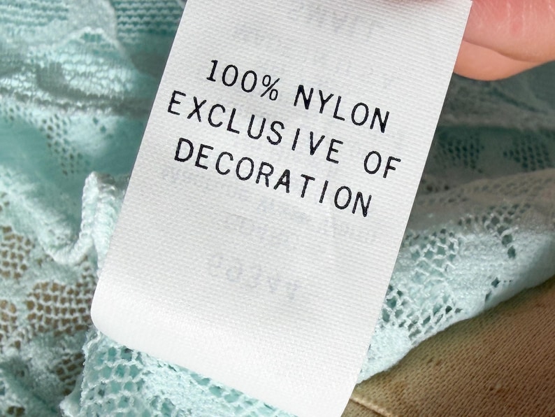 May include: A white fabric garment tag with black text that reads 100% NYLON EXCLUSIVE OF DECORATION, attached to a light mint green lace fabric with a detailed floral pattern. The tag is rectangular and positioned against the textured lace.