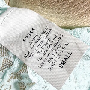 May include: A white fabric garment care label with black text reading 69344, CO4RC, Machine Wash Warm With Similar Colors, No Bleach, Tumble Dry Low, Cool Iron, If Needed, RN 79862, MADE IN U.S.A., and SMALL, attached to light blue lace fabric.