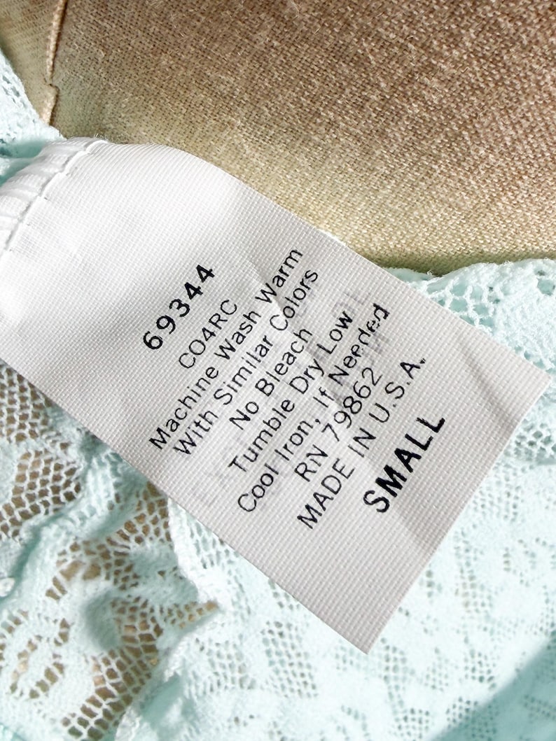 May include: A white fabric garment care label with black text reading 69344, CO4RC, Machine Wash Warm With Similar Colors, No Bleach, Tumble Dry Low, Cool Iron, If Needed, RN 79862, MADE IN U.S.A., and SMALL, attached to light blue lace fabric.