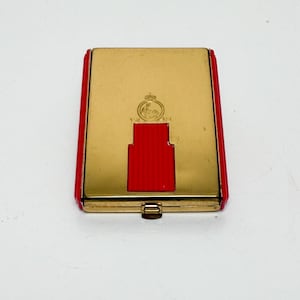 May include: A vintage gold-toned compact with red accents. The rectangular case features a red push-button clasp and red trim. A small emblem is visible on the gold surface. The compact is closed and appears to be in good condition.