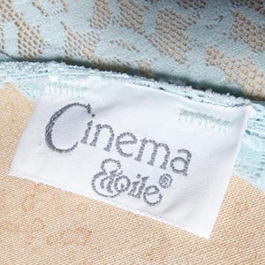 May include: A white fabric clothing label with the text Cinema Etoile in dark gray embroidery, attached to a light blue lace garment with a floral pattern. The label is rectangular and stitched onto the textured, pale blue lace fabric.
