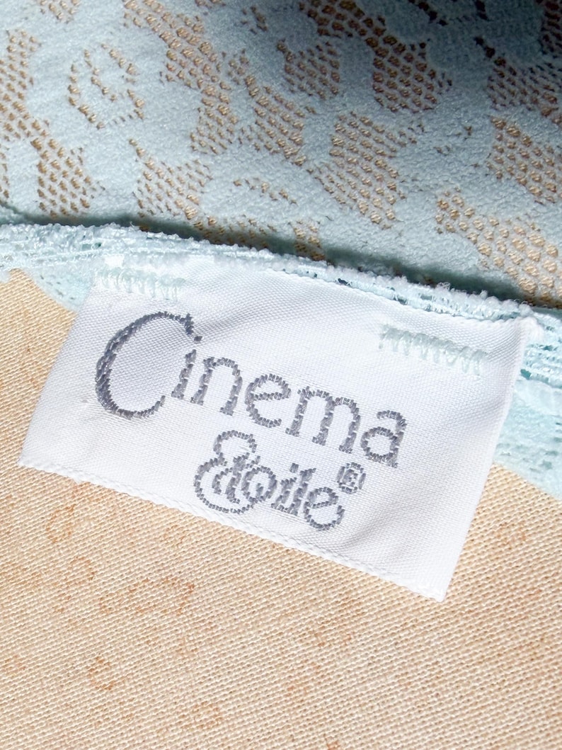 May include: A white fabric clothing label with the text Cinema Etoile in dark gray embroidery, attached to a light blue lace garment with a floral pattern. The label is rectangular and stitched onto the textured, pale blue lace fabric.