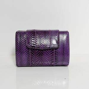 Puede incluir: Una cartera rectangular morada con un patrón de piel de serpiente texturizado. La cartera tiene un cierre de solapa con un broche y está hecha de un material brillante y duradero. La cartera es de un tono morado intenso.