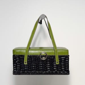 May include: A rectangular handbag featuring a black woven wicker base and a smooth olive green lid. The bag includes two olive green leather-like straps and a central silver-toned circular metal clasp on the front, providing a secure closure.
