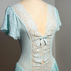 May include: A light blue nightgown featuring a deep V-neckline with intricate floral lace detailing. The garment includes short flutter sleeves, a central lace panel extending down the bodice, and a small satin ribbon bow at the center chest.