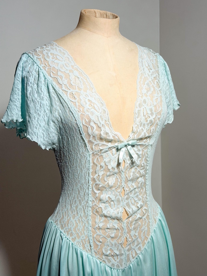 May include: A light blue nightgown featuring a deep V-neckline with intricate floral lace detailing. The garment includes short flutter sleeves, a central lace panel extending down the bodice, and a small satin ribbon bow at the center chest.