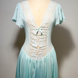May include: A light blue nightgown featuring a deep V-neckline with white lace trim, short flutter sleeves, and a central vertical lace panel accented by a small bow at the chest. The bodice is fitted with textured fabric, transitioning into a flowing skirt.