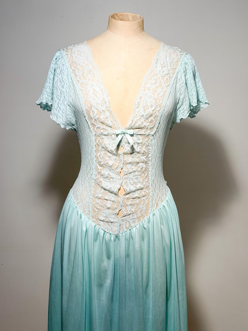 May include: A light blue nightgown featuring a deep V-neckline with white lace trim, short flutter sleeves, and a central vertical lace panel accented by a small bow at the chest. The bodice is fitted with textured fabric, transitioning into a flowing skirt.
