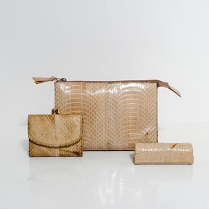 May include: A set of three beige leather accessories: a clutch with a zipper, a small wallet with a clasp, and a cylindrical case. The items have a reptile skin pattern and are arranged on a white surface.