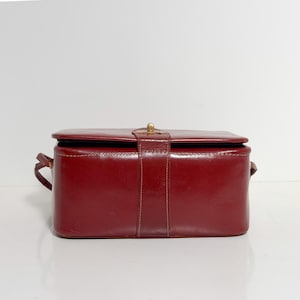 Puede incluir: Un bolso de mano rectangular de cuero burdeos con cierre de solapa y cierre dorado. El bolso tiene un diseño estructurado con un detalle de correa vertical y asas pequeñas a cada lado. El bolso mide aproximadamente 23 cm de ancho.