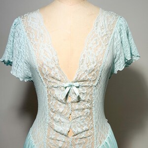 May include: A light blue nightgown featuring a deep V-neckline with intricate floral lace detailing. The bodice includes a small satin ribbon bow at the center, short flutter sleeves, and a smooth, flowing skirt made of a solid light blue fabric.