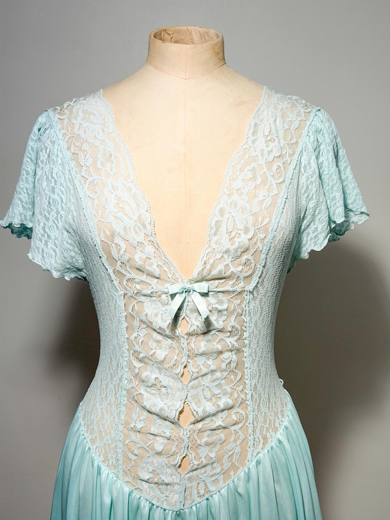 May include: A light blue nightgown featuring a deep V-neckline with intricate floral lace detailing. The bodice includes a small satin ribbon bow at the center, short flutter sleeves, and a smooth, flowing skirt made of a solid light blue fabric.