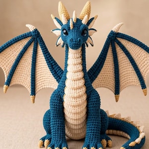 May include: A crocheted dragon plush toy featuring a deep blue body with a cream-colored underbelly, wings, and spikes. The creature has large dark eyes, pointed ears, and sharp-looking claws on its feet, all crafted with yarn.