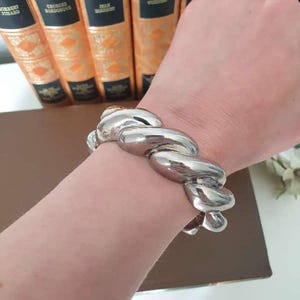 May include: A polished silver-tone cuff bracelet featuring a thick, twisted rope design. The bracelet has a high-shine, reflective metallic finish and a substantial, rounded structure that wraps around the wrist with a continuous, braided appearance.