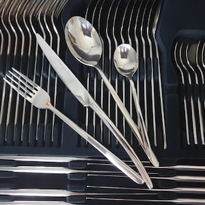 May include: A stainless steel flatware set arranged in a black molded tray. The set includes multiple forks, knives, and spoons with a polished finish. A single fork, knife, and two spoons are placed diagonally across the organized rows of cutlery.