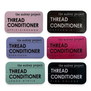 May include: Six rectangular metal tins of thread conditioner by the audrey project in pink, black, purple, magenta, mint green, and dark blue. Each tin features the text THREAD CONDITIONER and a specific scent name: Apple+Cinnamon, Peppermint, Bubblegum, Orange+Vanilla, Lemon Myrtle, and Pina Colada.