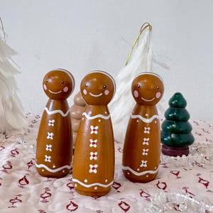 May include: Three brown wooden peg dolls painted to resemble gingerbread people. Each figure features a white icing-style wavy line pattern, small white flower accents, pink circular cheeks, and a simple painted smile on a round head.