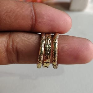 May include: Three thin gold-toned metal rings featuring intricate engraved patterns. Each ring displays a distinct textured design, including floral and geometric motifs, arranged side-by-side on a finger to showcase the detailed metalwork.