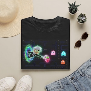 May include: A black short-sleeve t-shirt featuring a graphic print of a cartoon character emerging from a glowing green portal, reaching toward a pixelated strawberry. The design includes three pixelated ghosts in pink, blue, and orange colors.