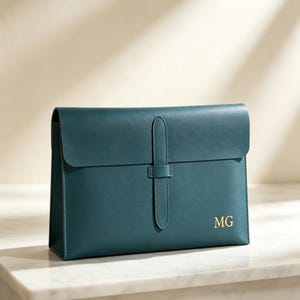 May include: A dark teal rectangular portfolio case with a smooth finish, featuring a vertical strap closure centred on the front flap and the gold-coloured embossed initials MG positioned in the bottom right corner of the main body.