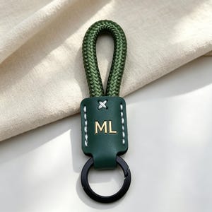 May include: A dark green keyring featuring a braided rope loop attached to a smooth leather tab. The leather tab is embossed with the gold letters ML and features white contrast stitching. A black metal ring is attached to the bottom.