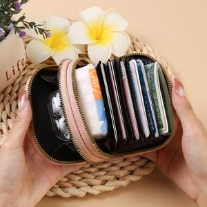 May include: A light pink accordion-style wallet with a gold-tone zipper closure. The interior features multiple black card slots and compartments containing various credit cards, paper currency, and a small clear case for wireless earbuds.