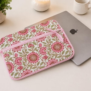 Puede incluir: Una funda acolchada para portátil con un estampado floral de flores rosas y hojas verdes sobre fondo blanco. La funda cuenta con un borde a rayas rosas y blancas, un bolsillo frontal con cremallera y tiradores de cremallera a juego a rayas.