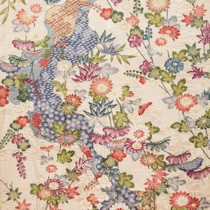 May include: A cream-colored fabric featuring a dense, intricate pattern of multi-colored flowers, including chrysanthemums and maple leaves, interspersed with winding, textured blue and tan streams decorated with geometric and floral motifs.