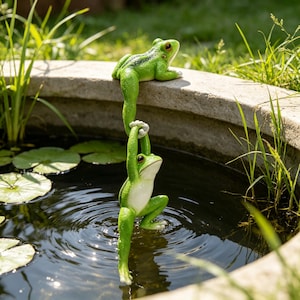 Puede incluir: Una escultura decorativa de jardín con dos ranas de color verde brillante. Una rana está sentada en el borde de un estanque de piedra, mientras que la otra cuelga debajo, agarrada a la pata de la primera. Ambas tienen piel lisa y ojos oscuros.