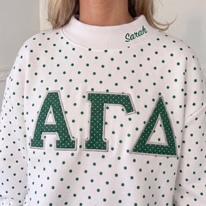 May include: A white sweatshirt featuring a green polka dot pattern and a ribbed mock neck collar. The name Sarah is embroidered in green on the collar. Large Greek letters Alpha, Gamma, and Delta are appliqued on the chest in green with white dots.