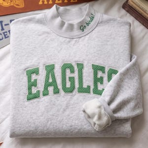 Könnte beinhalten: Ein hellgrau meliertes Sweatshirt mit Stehkragen und dem Wort EAGLES in großen grünen Buchstaben mit weißen Punkten auf der Brust. Der Kragen ist mit dem grünen Sticktext Go birds! versehen und die Innenseite ist mit weichem Fleece gefüttert.
