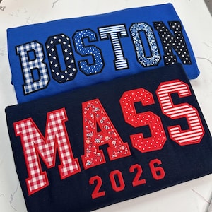 May include: A blue sweatshirt with the word BOSTON in patterned appliqué letters and a navy sweatshirt with the word MASS and the year 2026 in red patterned appliqué letters. The letters feature various prints including gingham, stars, and floral.