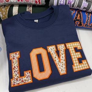 Peut inclure: Un sweat-shirt bleu marine à col rond arborant le mot LOVE en grandes lettres style universitaire. Chaque lettre est soulignée d'orange vif et remplie d'un tissu à motifs différent, incluant des fleurs, des rayures et un vichy jaune.