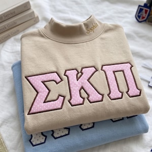 Puede incluir: Una sudadera beige de cuello alto con las letras griegas Sigma Kappa Pi en rosa con lunares blancos y un borde marrón oscuro. El nombre Sophia está bordado en hilo dorado en el cuello. Debajo hay una sudadera de color azul claro.
