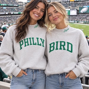 Peut inclure: Deux sweat-shirts gris clair à col rond avec des lettres brodées en vert. L'un affiche le mot PHILLY dans une police arquée, tandis que l'autre affiche le mot BIRD. Les deux portent le nom Sophia brodé sur le col.