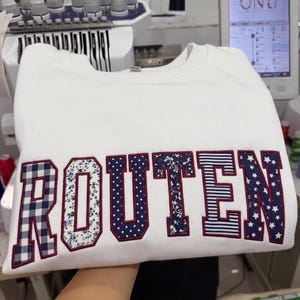 Puede incluir: Una sudadera blanca de cuello redondo con la palabra ROUTEN en letras grandes de estilo bloque. Cada letra está rellena con una tela diferente de estampado azul marino y blanco, incluyendo cuadros vichy, flores, lunares y estrellas, bordeada en granate.