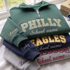 Pode incluir: Uma pilha de três camisolas com fecho de um quarto em verde, azul-marinho e cinzento mesclado. Cada uma apresenta texto bordado incluindo PHILLY, EAGLES e BULLDOGS com padrões axadrezados, além de School name e nomes como Lucas e Emily.