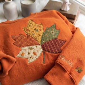 Könnte beinhalten: Ein orangefarbenes Sweatshirt mit einer großen, gesteppten Ahornblatt-Applikation auf der Vorderseite. Das Blatt besteht aus verschiedenen Stoffstücken mit gelben, grünen und braunen Mustern. Auf dem Ärmel ist der Name Charlotte in Schreibschrift gestickt.