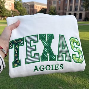 May include: A white crewneck sweatshirt featuring the word TEXAS in large, block-style letters with varied green and white patterns, including floral, striped, polka dot, and gingham prints. The word AGGIES is embroidered in green below the letters.