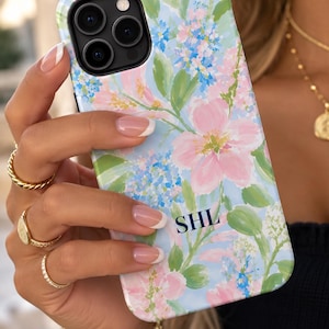 May include: A smartphone case featuring a light blue background with a painted floral pattern in shades of pink, green, and blue. The design includes visible brushstroke textures and the black capital letters SHL printed near the bottom center.