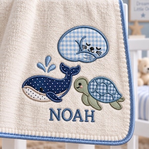 Puede incluir: Una toalla de color crema con un borde ribeteado en azul que presenta diseños bordados de una foca durmiendo con patrón de cuadros azules, una ballena azul oscuro con lunares y chorro de agua, una tortuga verde con caparazón de cuadros y el nombre NOAH.