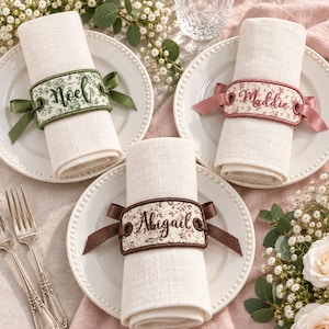 May include: Three off-white linen napkins rolled and secured with personalised fabric napkin rings. Each ring features a floral pattern, a satin ribbon tie, and embroidered names: Noel in green, Maddie in pink, and Abigail in brown.