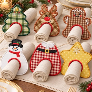 May include: A set of six embroidered Christmas napkin rings featuring a green plaid tree, a brown plaid reindeer, a brown gingerbread person, a white snowman with a black hat, a red and white plaid Santa suit, and a yellow star with snowflakes.