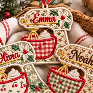 Puede incluir: Tres adornos bordados con forma de batidora de pie con los nombres Emma, Olivia y Noah. Cada adorno muestra una batidora con un bol de nata montada, decorado con flores de pascua, cerezas y patrones de cuadros, con una cinta roja.